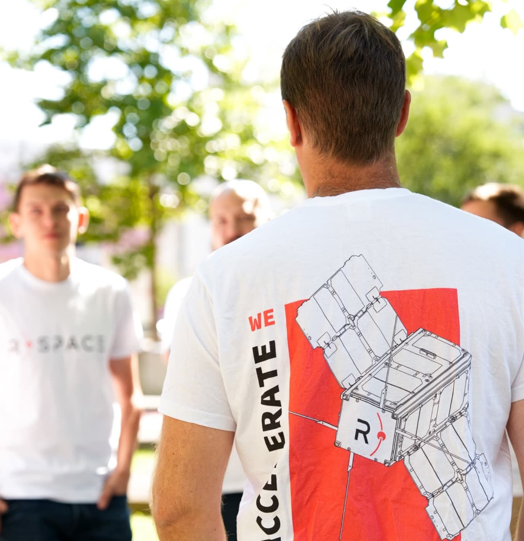 Group of people wearing R-Space T-shirts outdoors, with one shirt prominently showing a CubeSat satellite illustration and the text ‘We Accelerate.