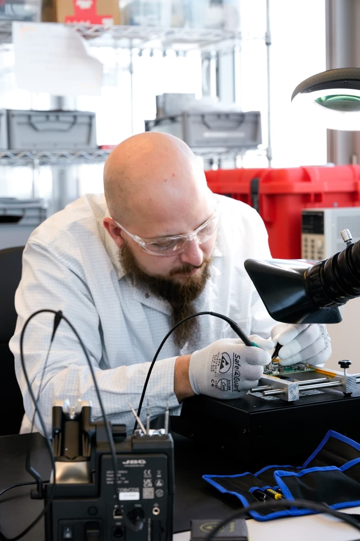 Engineer wearing safety glasses and gloves soldering or testing an electronic circuit board in a laboratory workspace.