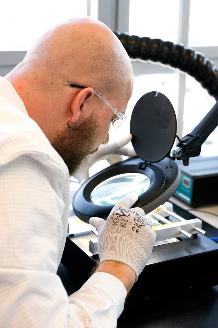 Engineer wearing gloves and safety glasses inspecting small electronic components through a magnifying lamp in a laboratory.