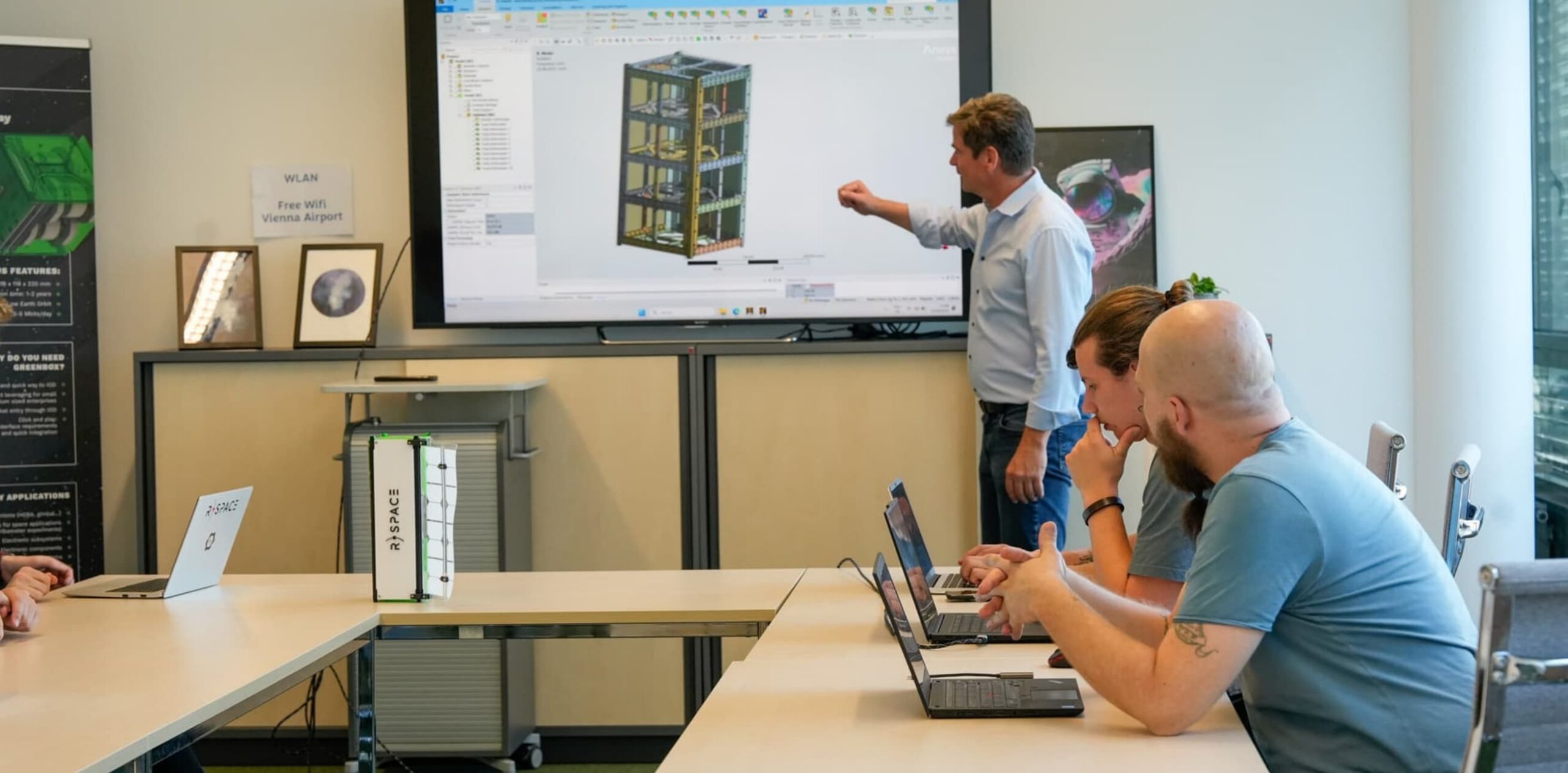 Team meeting at R-Space with a presenter showing a 3D model of a satellite or CubeSat on a large screen while colleagues with laptops discuss and a physical satellite prototype sits on the table.