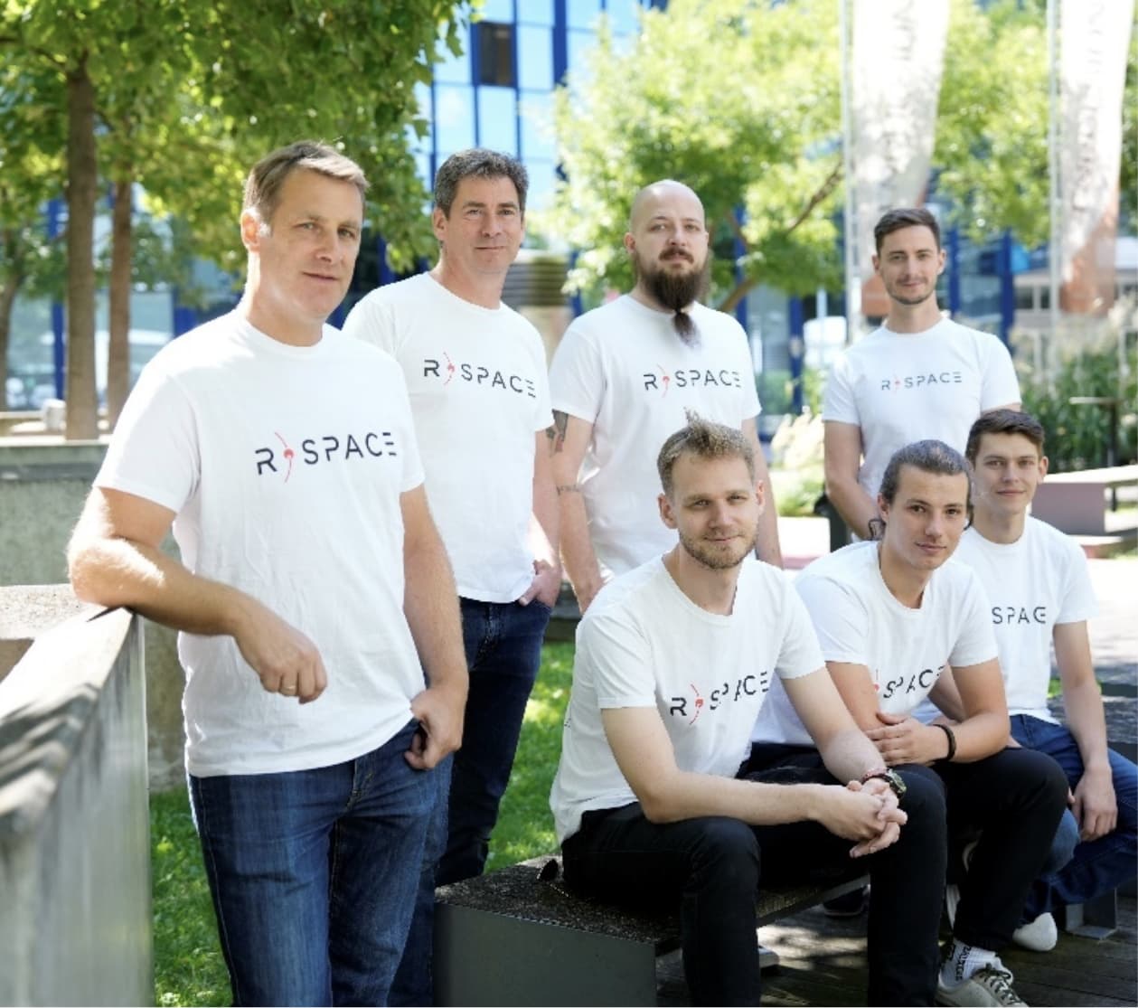 Group of seven people wearing matching R-Space T-shirts posing outdoors in a modern campus or office park setting